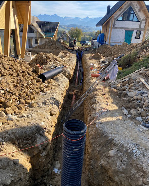 Głęboki wykop z ułożonymi rurami instalacyjnymi w ziemi, widok z góry, w tle budynki w trakcie budowy i krajobraz górski.