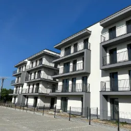 Nowoczesny budynek wielorodzinny z balkonami, wykończony w bieli i szarości, z widocznym parkingiem z kostki betonowej i ogrodzeniem panelowym.