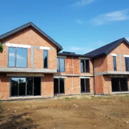 Dwukondygnacyjny, murowany dom w stanie surowym otwartym z zamontowanymi oknami, widoczne niedokończone ściany z czerwonej cegły i czarny dach, otoczony ziemią i zielenią.