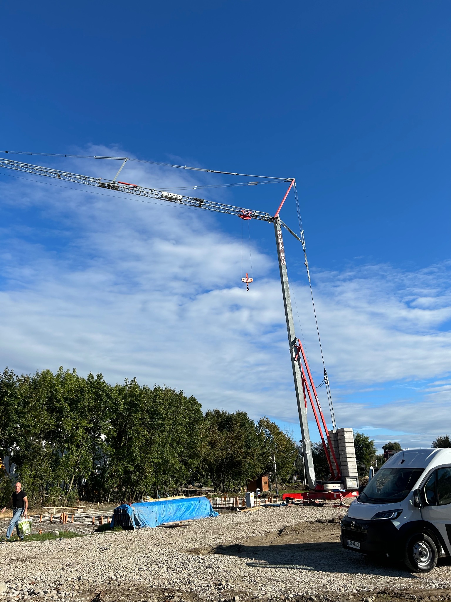 Budowa w toku: żuraw wieżowy Huber Bau na tle błękitnego nieba z chmurami. Na pierwszym planie teren budowy z fundamentami i zaparkowany biały van.