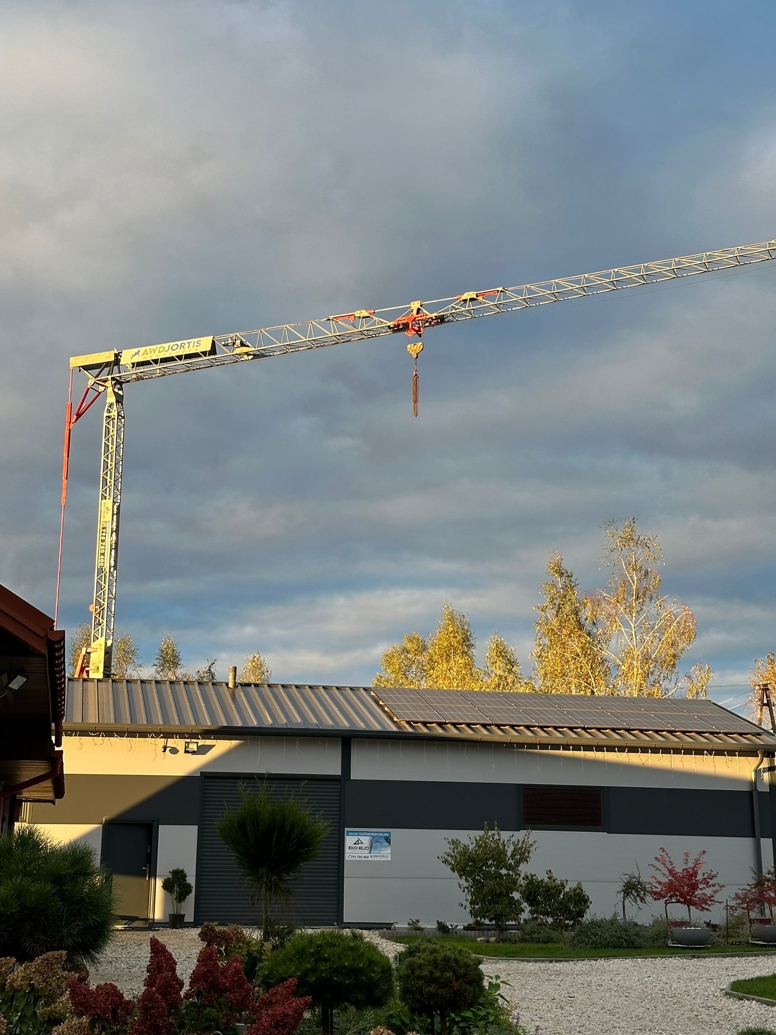 Żuraw budowlany na tle pochmurnego nieba, widoczny budynek z panelami słonecznymi na dachu i zagospodarowany teren wokół. Ujęcie z dołu, wieczorne światło.