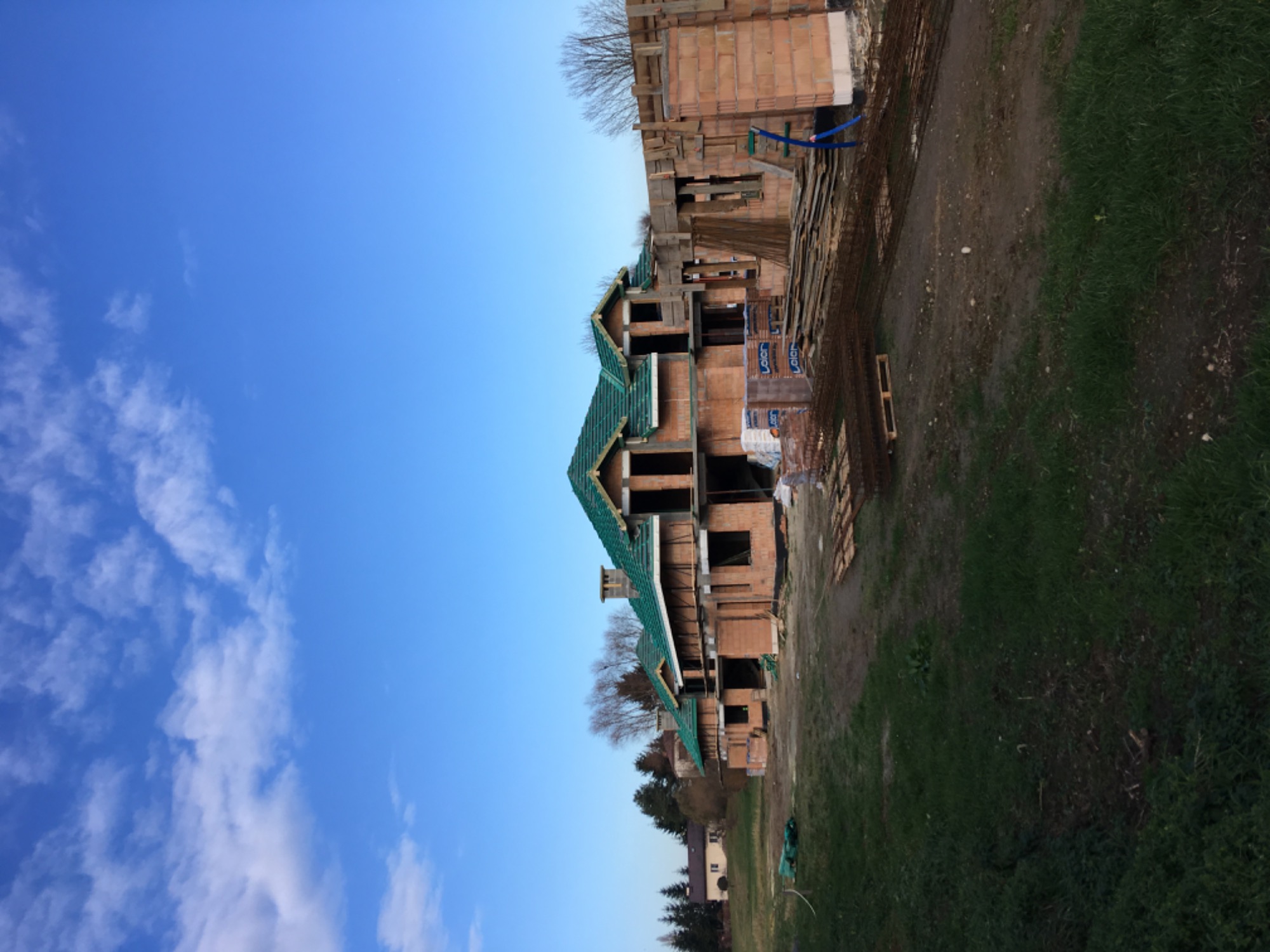 Niedokończony dom z cegły z zielonym dachem na tle błękitnego nieba i zieleni, widoczne materiały budowlane i zbrojenia.