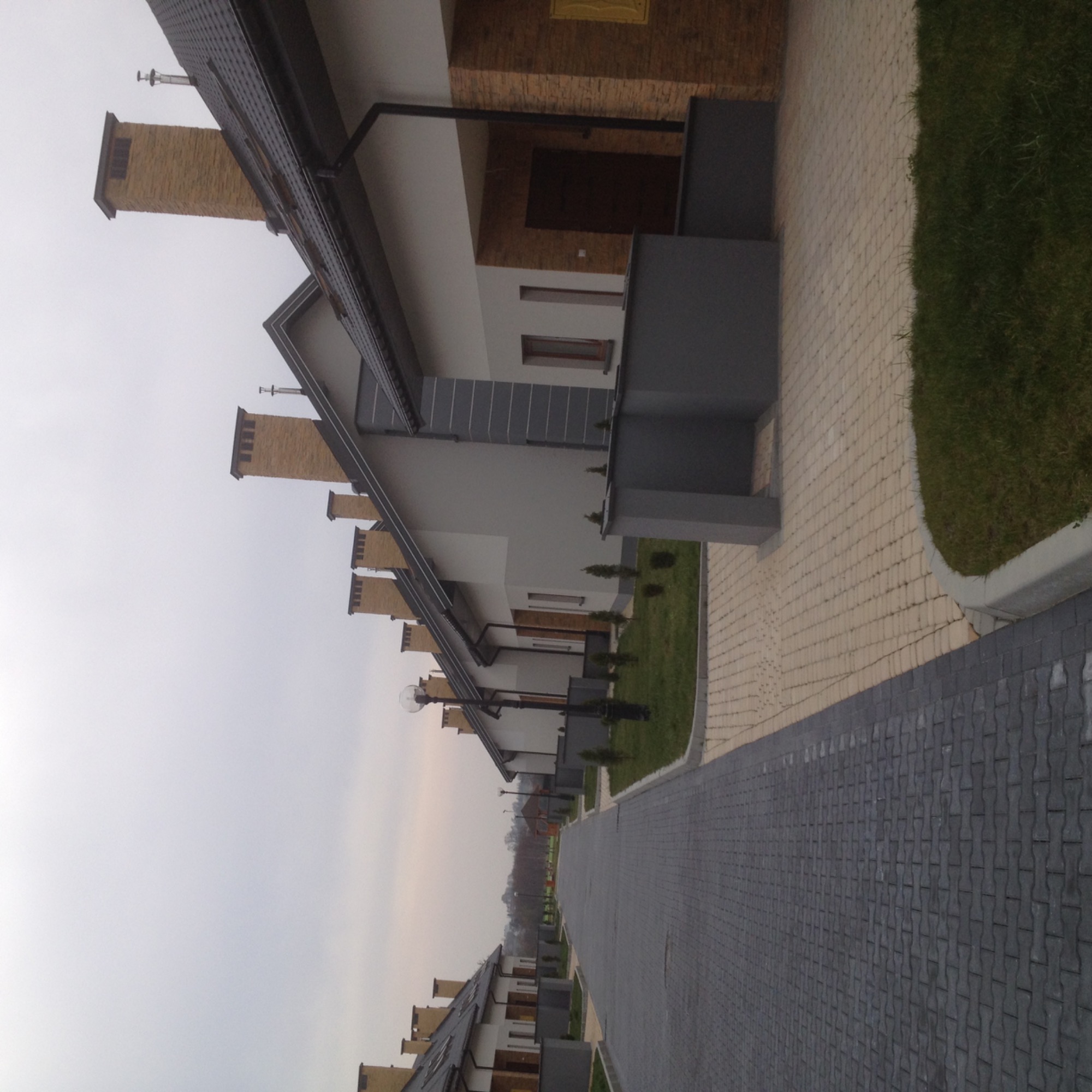 Nowoczesne osiedle domów jednorodzinnych z ceglanymi kominami, szarymi elementami elewacji i kostką brukową na podjeździe, widok z poziomu chodnika w pochmurny dzień.