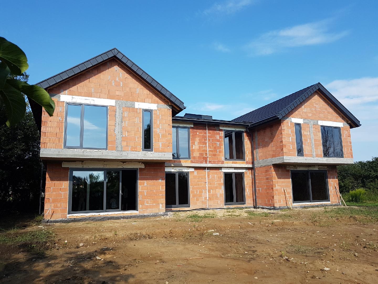 Dwukondygnacyjny dom murowany w trakcie budowy, z zamontowanymi oknami i dachem, widoczne niedokończone ściany z czerwonej cegły i teren budowy.