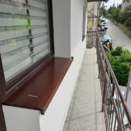 Biała elewacja budynku z widokiem z balkonu na ulicę, brązowe okno z żaluzjami, lekko ziarnista faktura tynku.