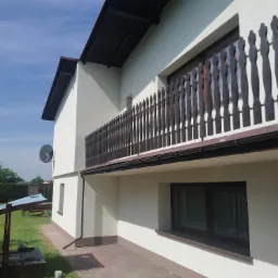 Świeżo pomalowana, jasna elewacja dwupiętrowego domu z ciemnymi elementami wykończeniowymi, takimi jak balustrady balkonowe i rynny, widoczna z poziomu chodnika z kostki, z wiadrem farby w rogu kadru.