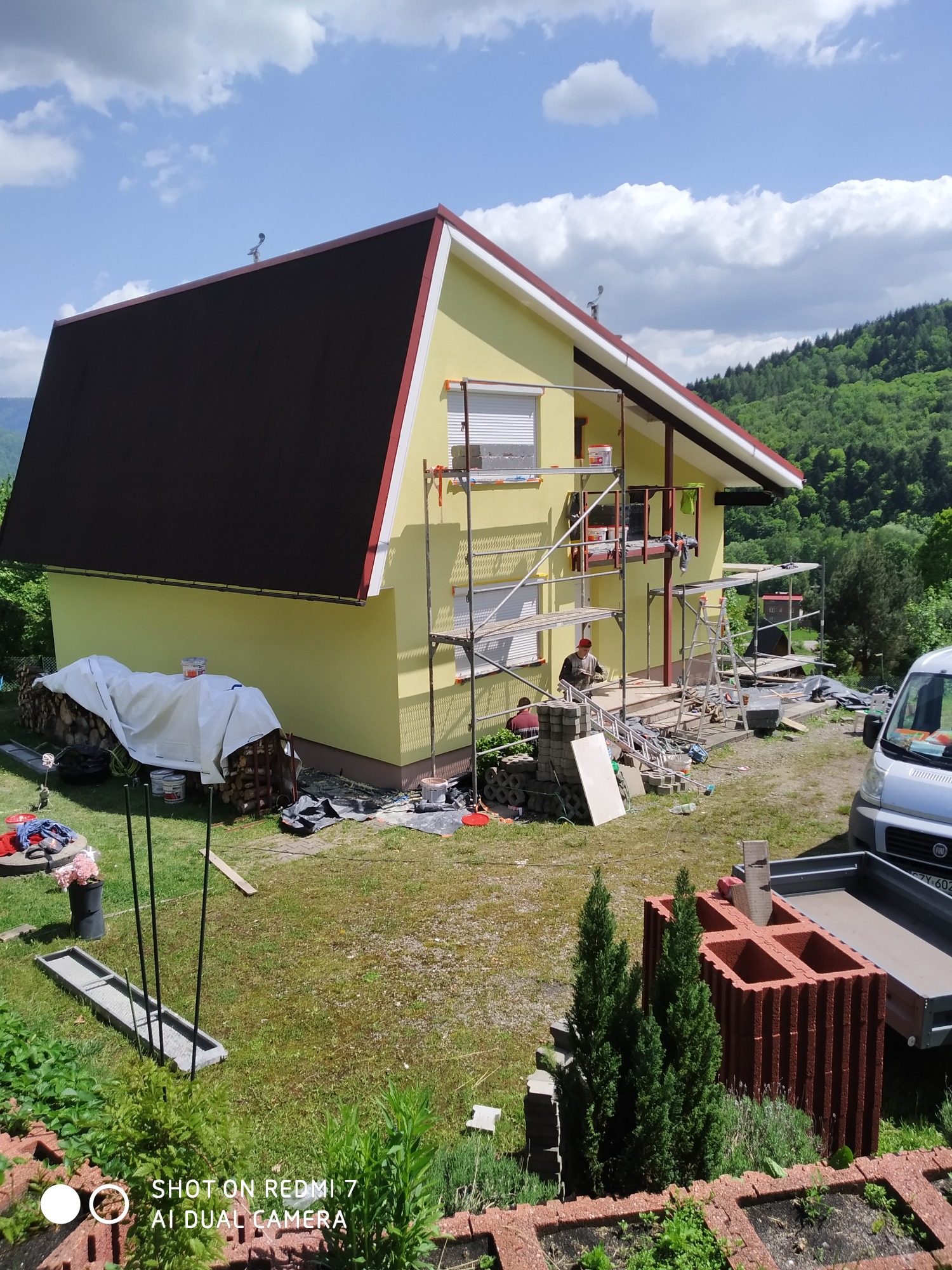 Żółty dom jednorodzinny w trakcie prac elewacyjnych, widoczne rusztowania, pracownicy, materiały budowlane i samochód dostawczy na tle zielonego wzgórza.