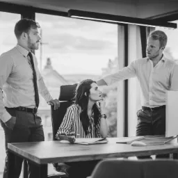 Czarno-białe zdjęcie przedstawia troje osób w nowoczesnym biurze: dwóch mężczyzn w koszulach i spodniach oraz kobietę w bluzce w paski siedzącą za biurkiem z dokumentami i komputerem. Jeden...