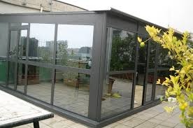 Zabudowa tarasu z szarymi profilami, widok na miasto zza szyb, fragment blatu stołu w lewym dolnym rogu, roślina z żółtymi liśćmi po prawej.