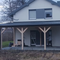 JT BUD - Nowoczesny dom z białymi ścianami i szarymi elementami, w trakcie budowy drewnianej konstrukcji zadaszenia tarasu, widoczna osoba pracująca przy drzwiach wejściowych.