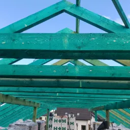 Konstrukcja więźby dachowej zaimpregnowana na zielono, widok od dołu z widocznymi belkami, łatami, membraną oraz materiałami budowlanymi na dachu w tle zabudowa jednorodzinna.