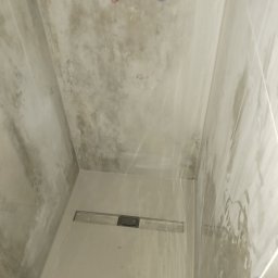 JT BUD - Wnętrze kabiny prysznicowej z odpływem liniowym w posadzce i przygotowanymi przyłączami wodnymi na ścianie, wykończonej płytkami imitującymi beton.