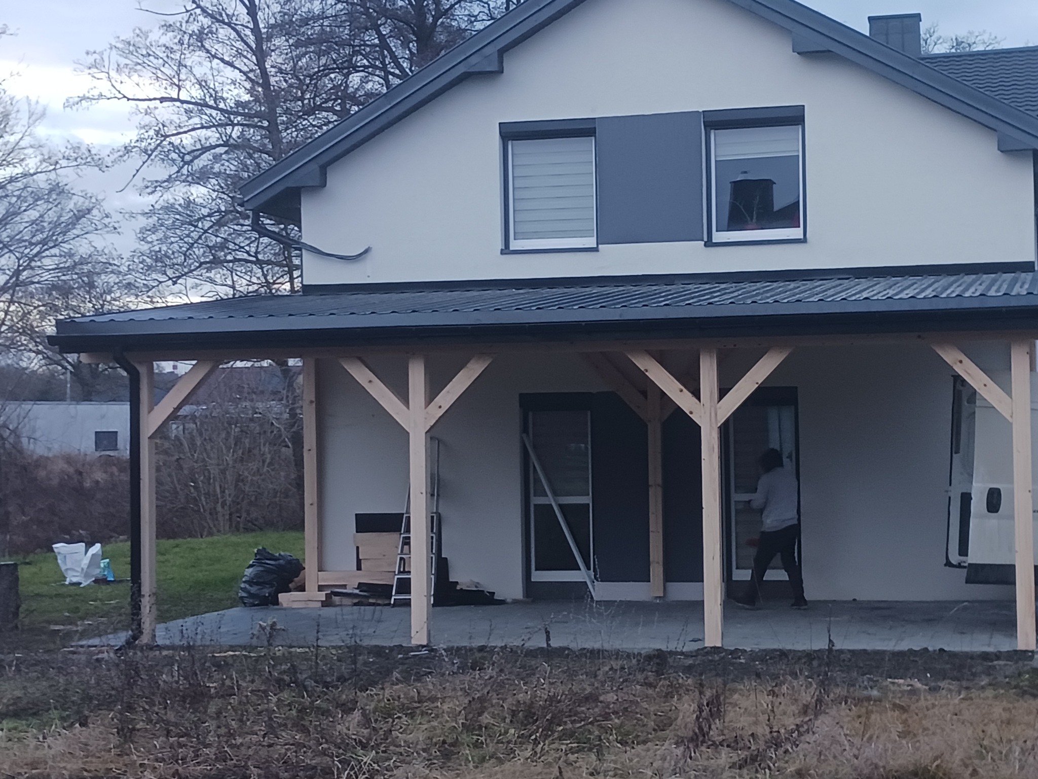 Nowoczesny dom z białymi ścianami i szarymi elementami, w trakcie budowy drewnianej konstrukcji zadaszenia tarasu, widoczna osoba pracująca przy drzwiach wejściowych.