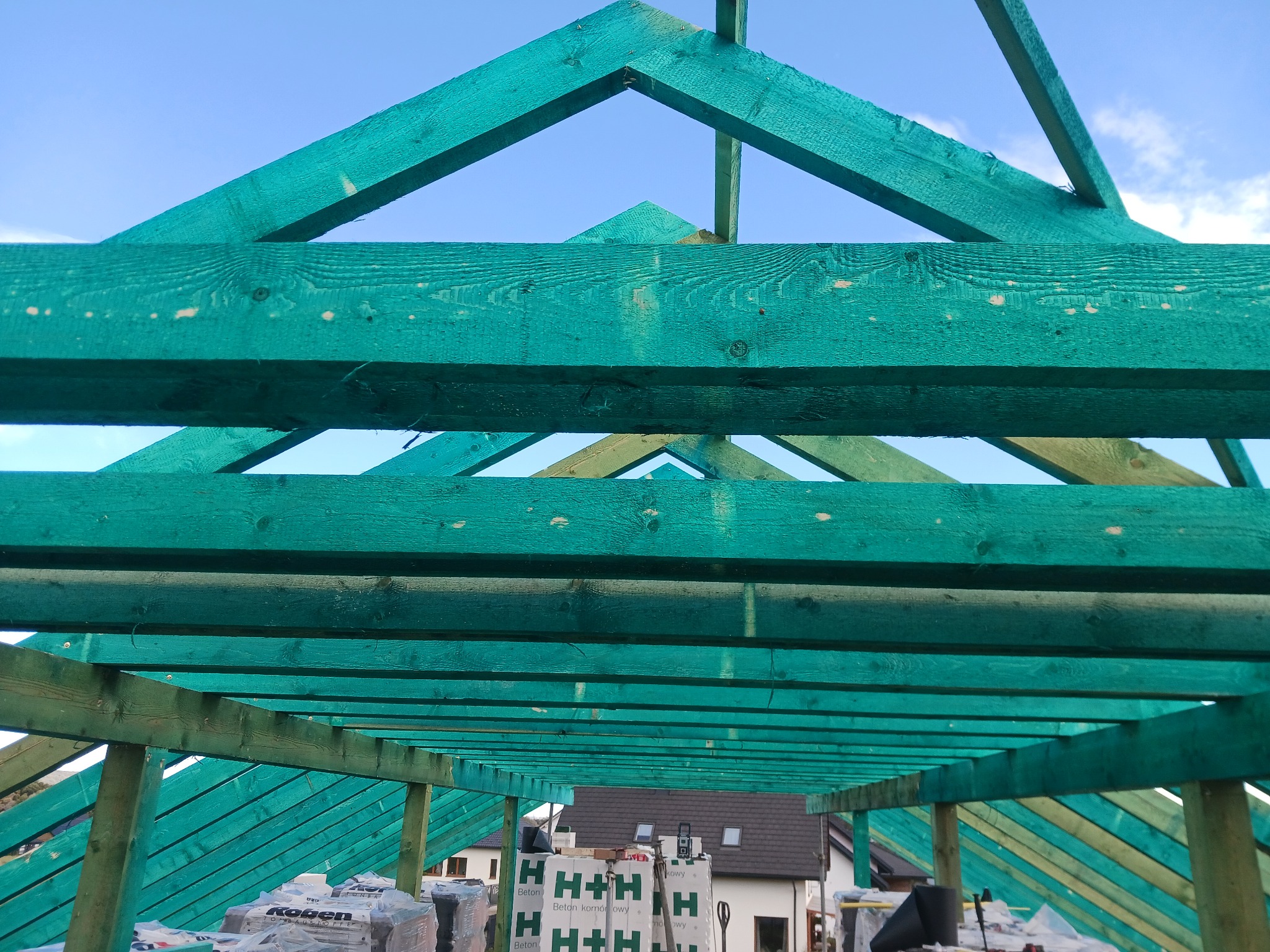 Konstrukcja więźby dachowej zaimpregnowana na zielono, widok od dołu z widocznymi belkami, łatami, membraną oraz materiałami budowlanymi na dachu w tle zabudowa jednorodzinna.