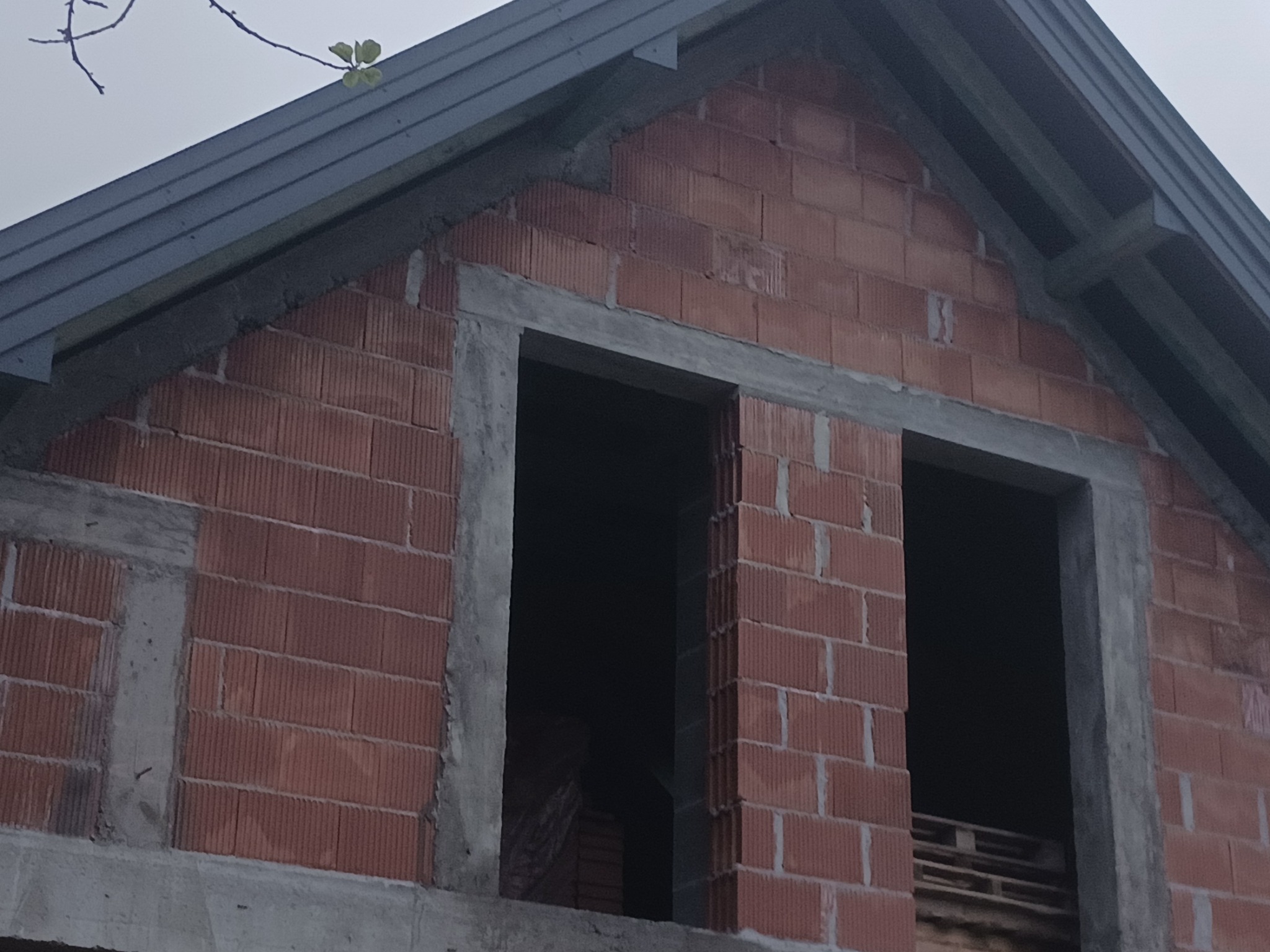 Surowy stan budynku z czerwonej cegły, widoczne otwory okienne z szarymi nadprożami betonowymi, fragment dachu z szarą blachą i gałązka z zielonymi listkami w górnym rogu.