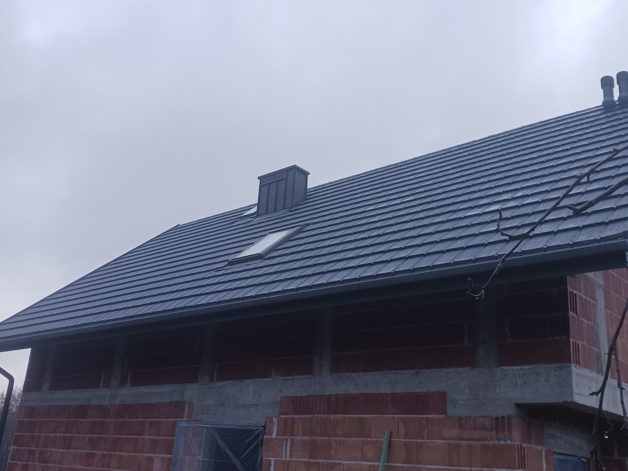 Szary dach z dachówki ceramicznej na budynku w trakcie budowy, widoczne okno dachowe i komin wentylacyjny.