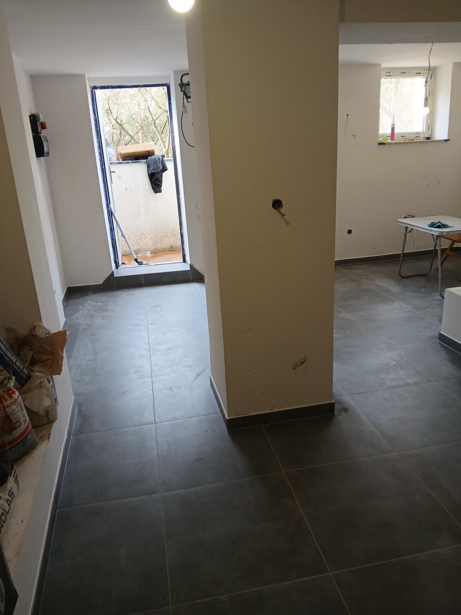 Wnętrze w trakcie remontu: ułożone ciemnoszare płytki podłogowe, widoczne worki z zaprawą murarską, kolumna nośna z nieotynkowaną ścianą, okno i drzwi balkonowe w tle.