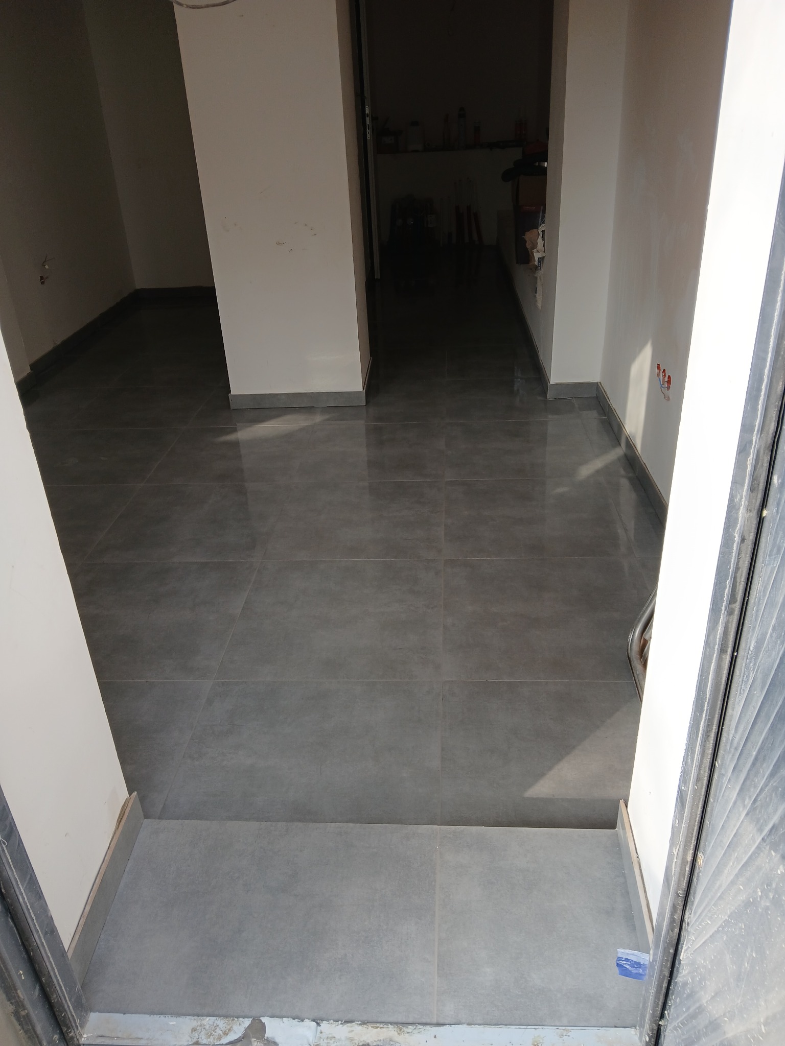 Ułożona, szara, kwadratowa terakota w nowym pomieszczeniu z białymi ścianami i widocznym progiem wejściowym oraz fragmentem otwartych drzwi.