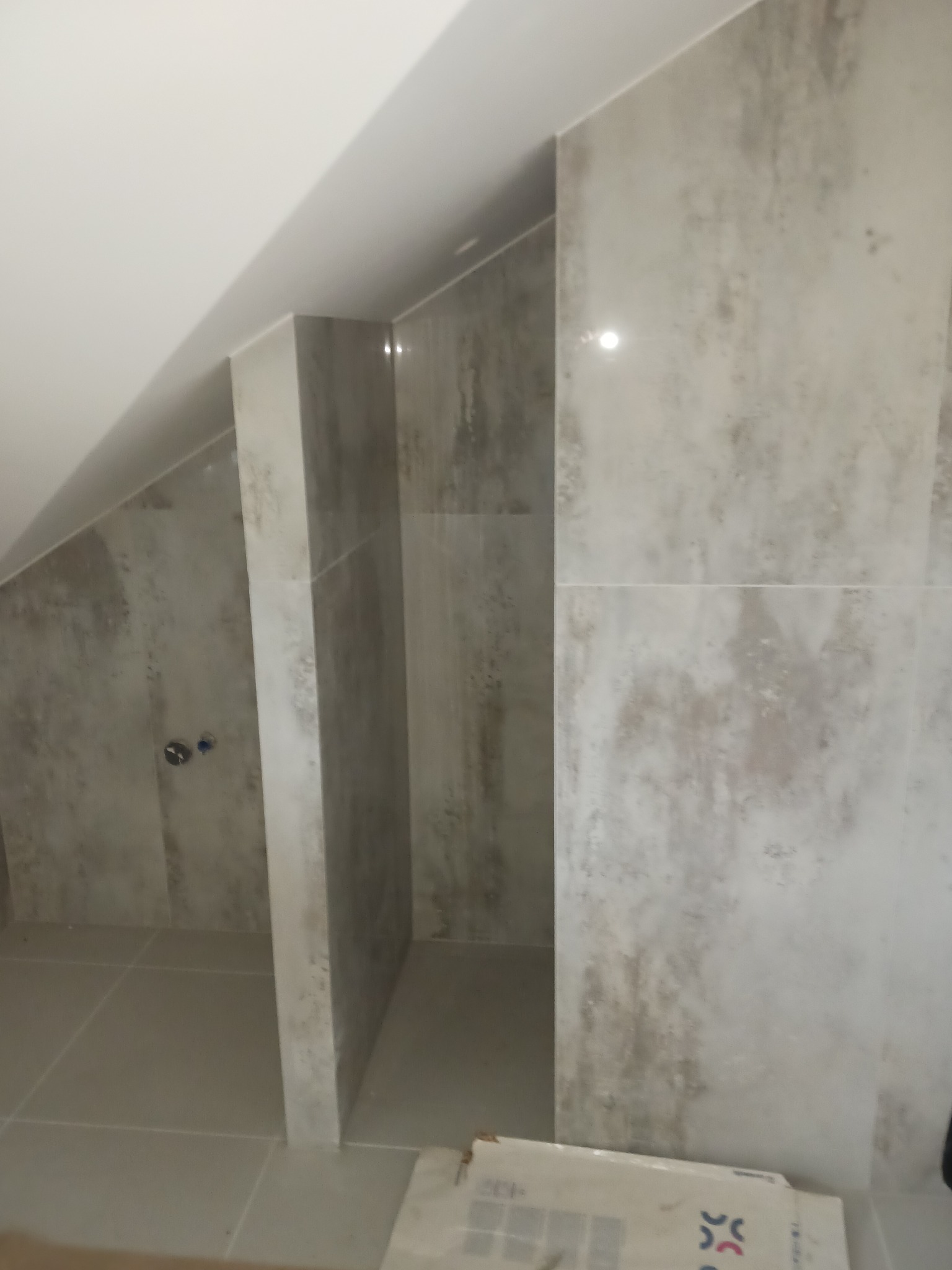 Wykończona łazienka na poddaszu z szarymi płytkami imitującymi beton, widoczny fragment kabiny prysznicowej i karton płytek w rogu.