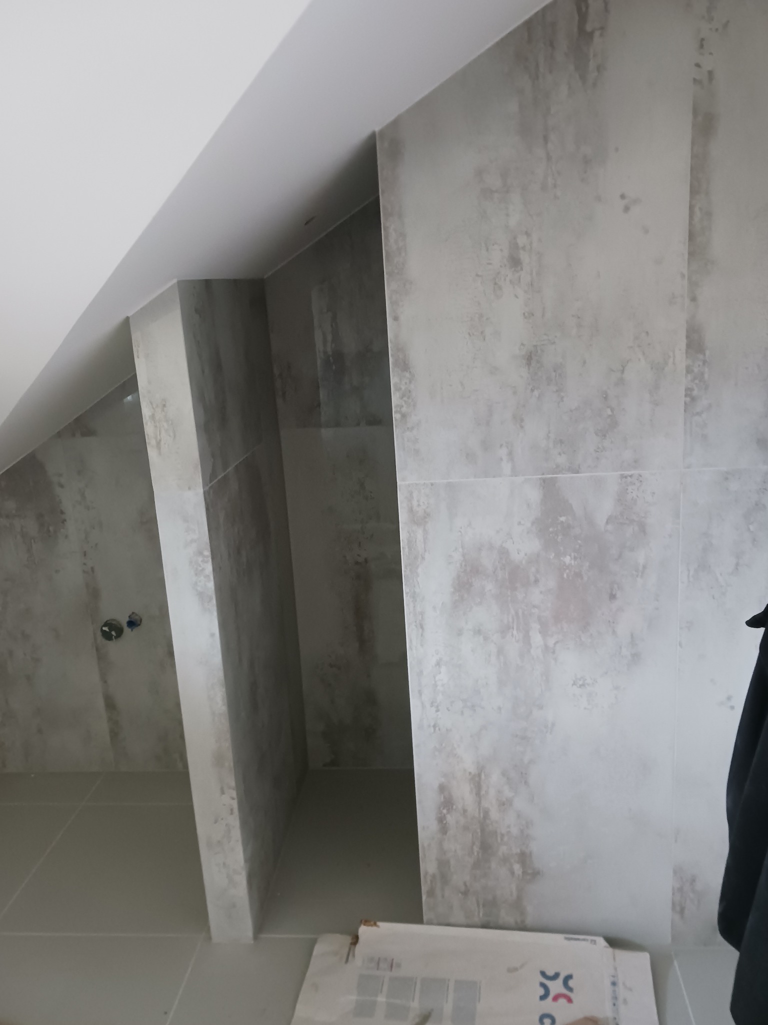 Wykończona łazienka na poddaszu ze ścianami i podłogą wyłożonymi płytkami w odcieniach szarości, widok na kabinę prysznicową z zamontowaną armaturą, fragment kartonu z płytkami w rogu.