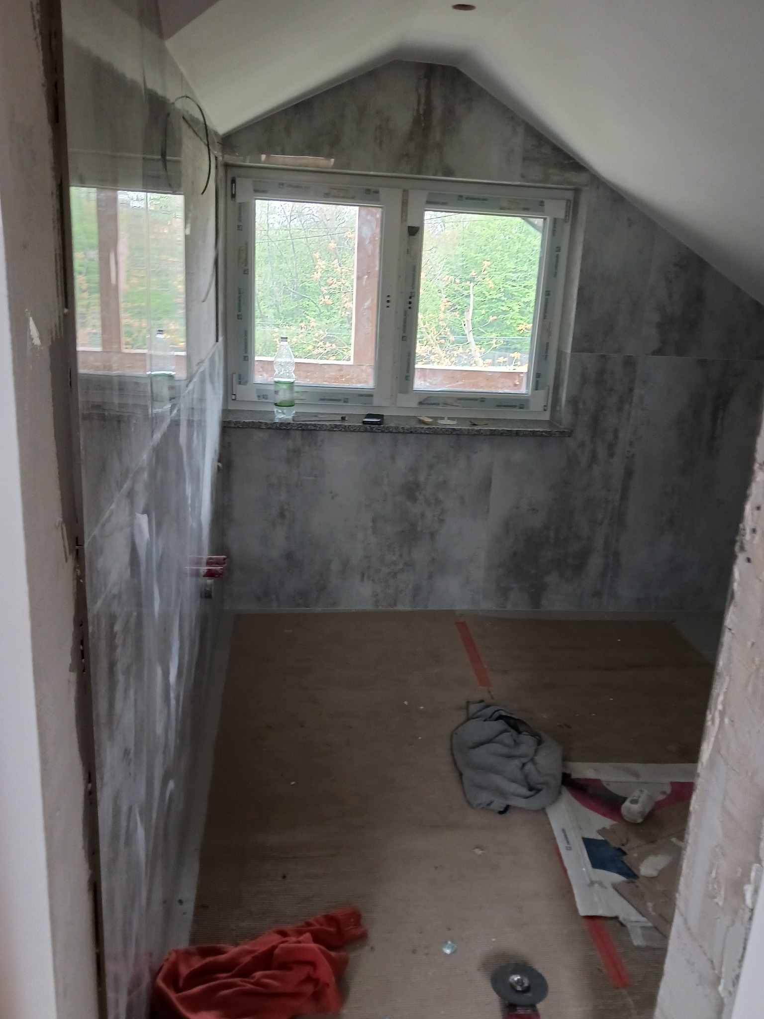 Łazienka na poddaszu w trakcie remontu, widoczne płytki imitujące beton, okno dachowe z widokiem na drzewa, butelka wody na parapecie, narzędzia i materiały budowlane na podłodze, ściany w trakcie...