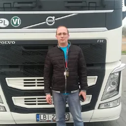 Kierowca w okularach i kurtce stoi na tle białej ciężarówki Volvo FH z polską rejestracją LBI, w tle widoczny fragment parkingu.