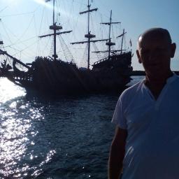 Non stop car - Sylwetka mężczyzny w białej koszulce na tle repliki żaglowca pirackiego na wodzie z refleksami słońca.