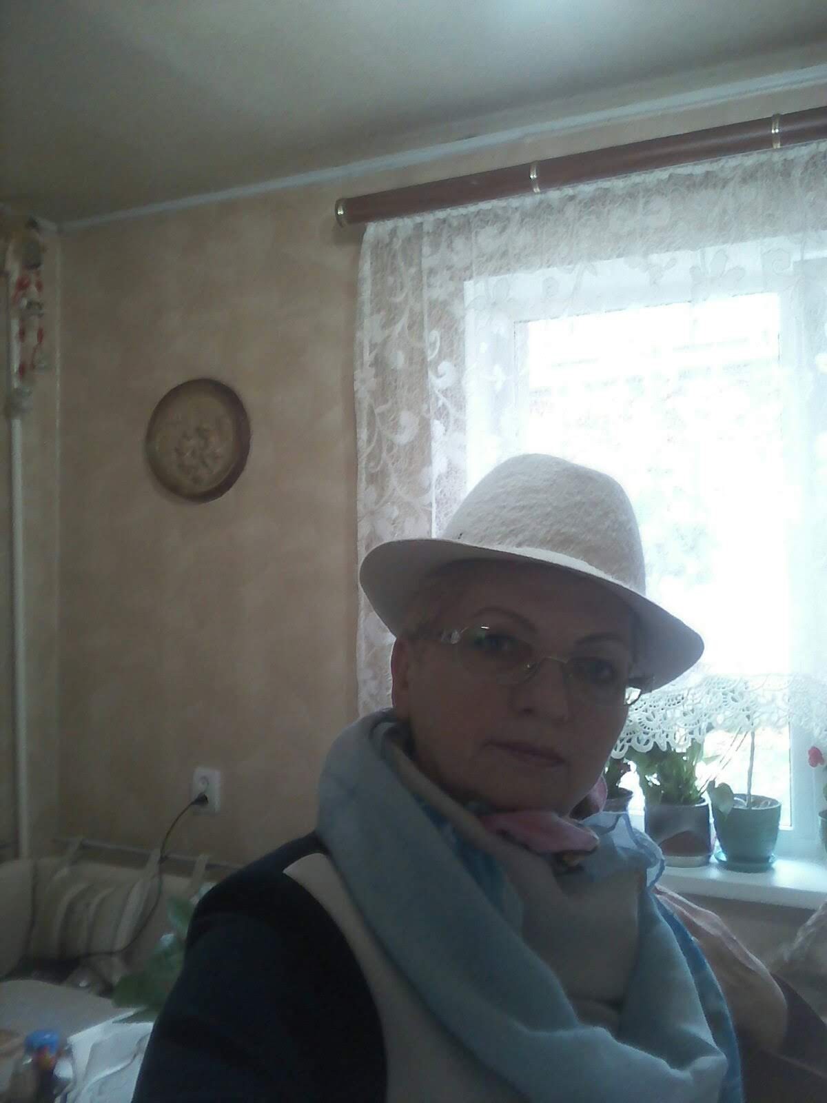 Portret kobiety w kapeluszu i szalu na tle okna z koronkową firanką i ścianą z dekoracyjnym talerzem.