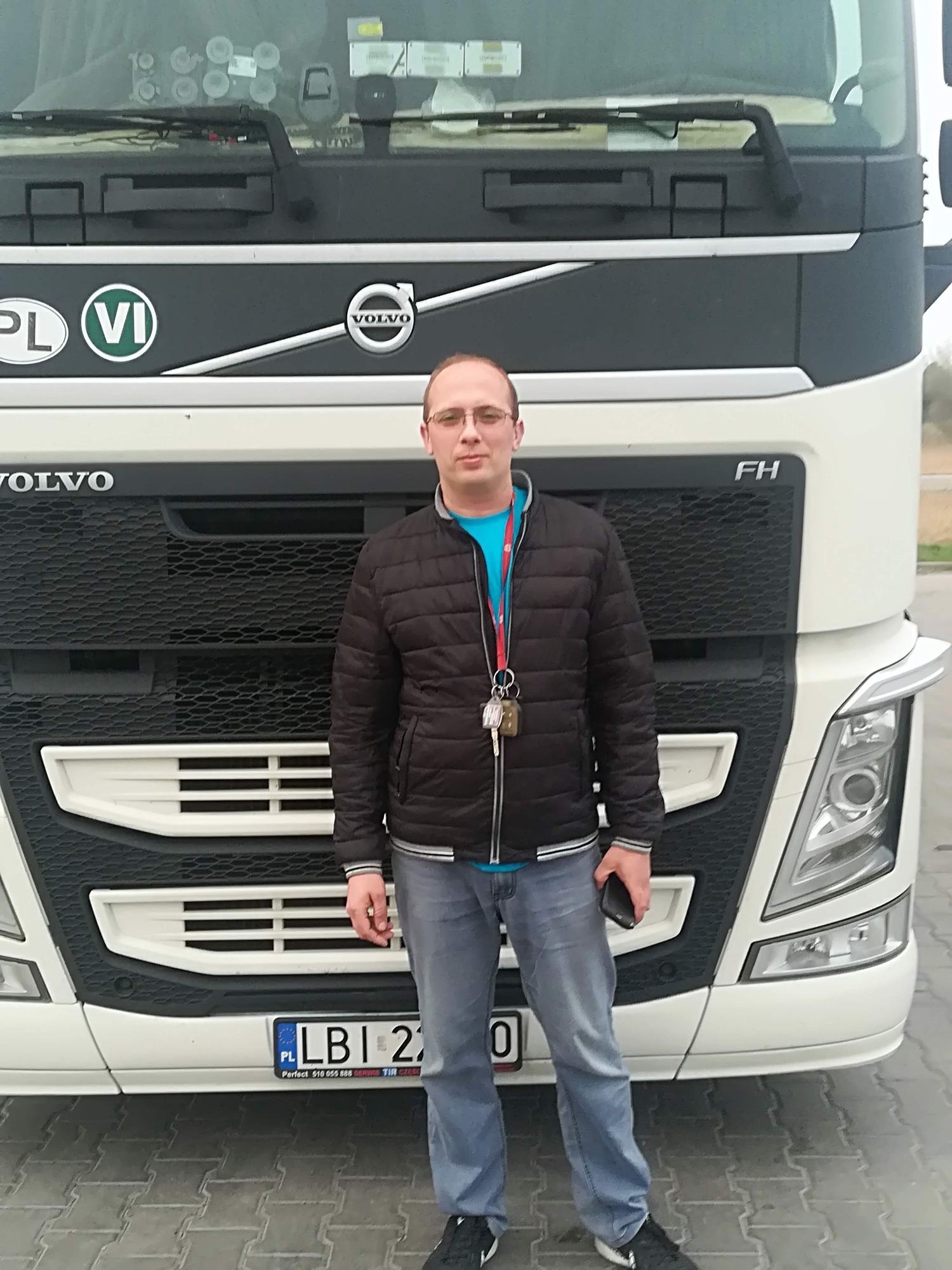 Kierowca w okularach i kurtce stoi na tle białej ciężarówki Volvo FH z polską rejestracją LBI, w tle widoczny fragment parkingu.