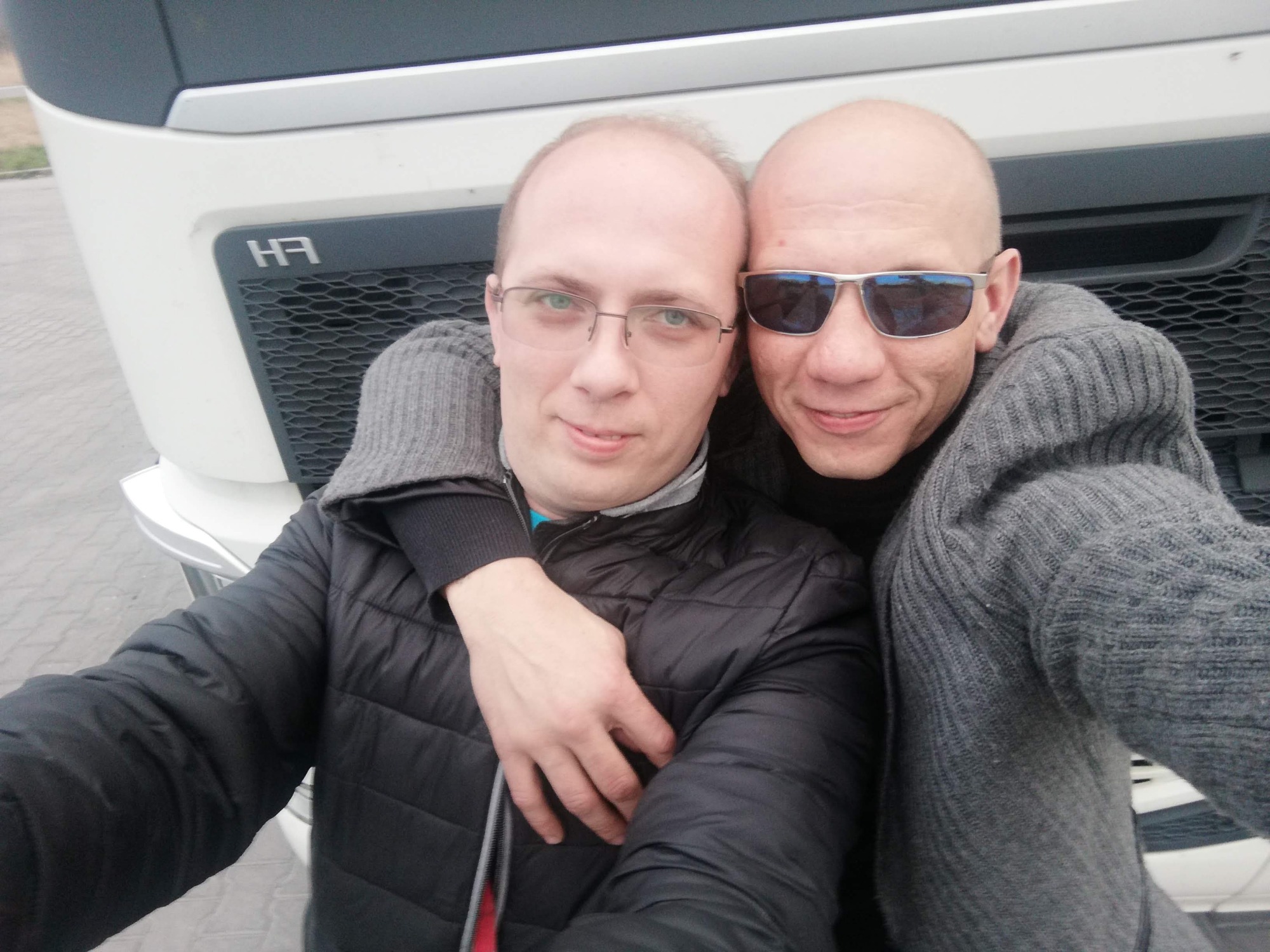 Dwaj mężczyźni obejmujący się na tle białej kabiny ciężarówki z widocznym logo 'H3' na grillu.