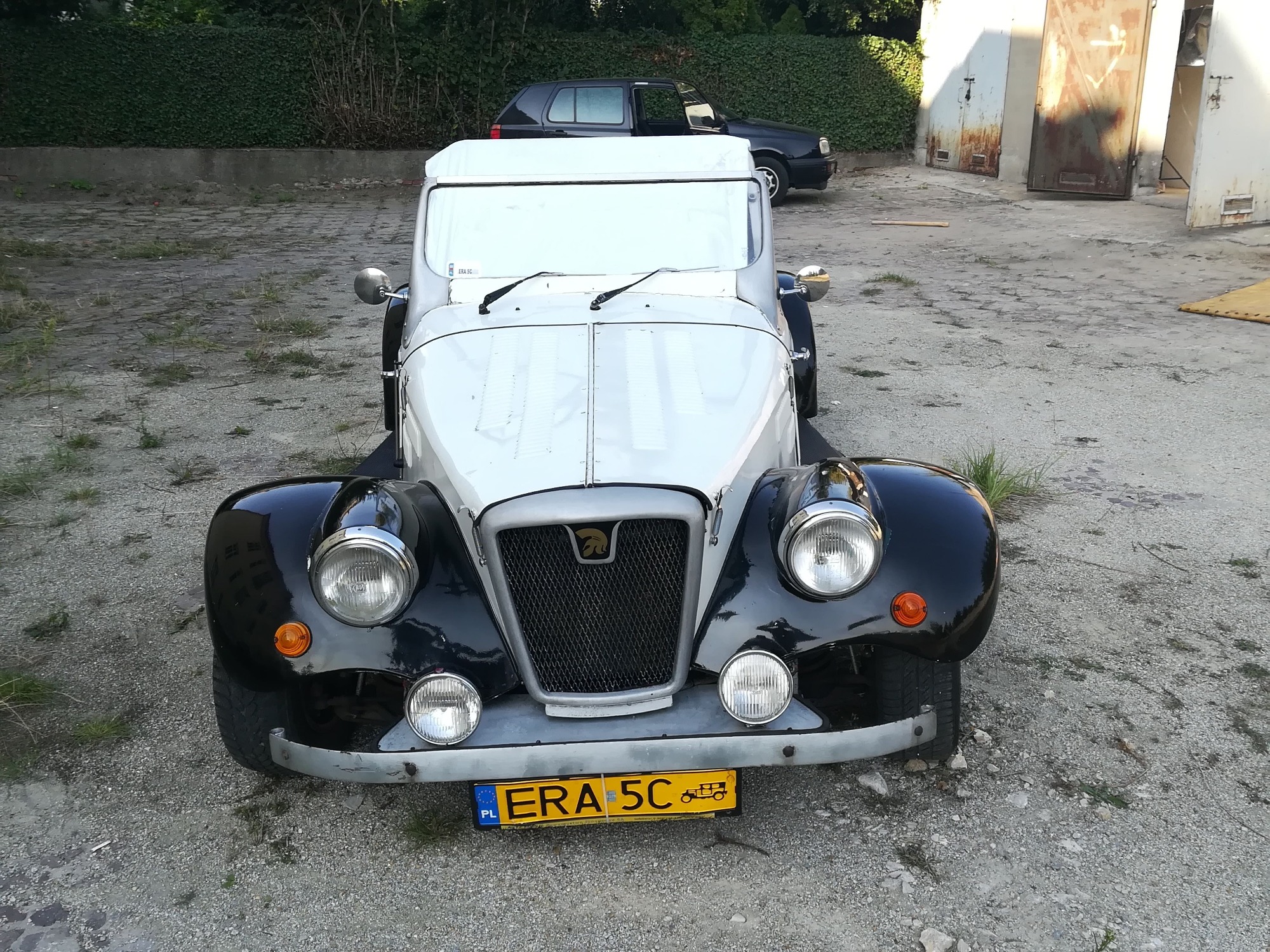 Przód białego, stylizowanego samochodu retro z czarnymi błotnikami i tablicą rejestracyjną ERA 5C, stojącego na szarym, żwirowym podłożu, z zielonymi krzewami w tle