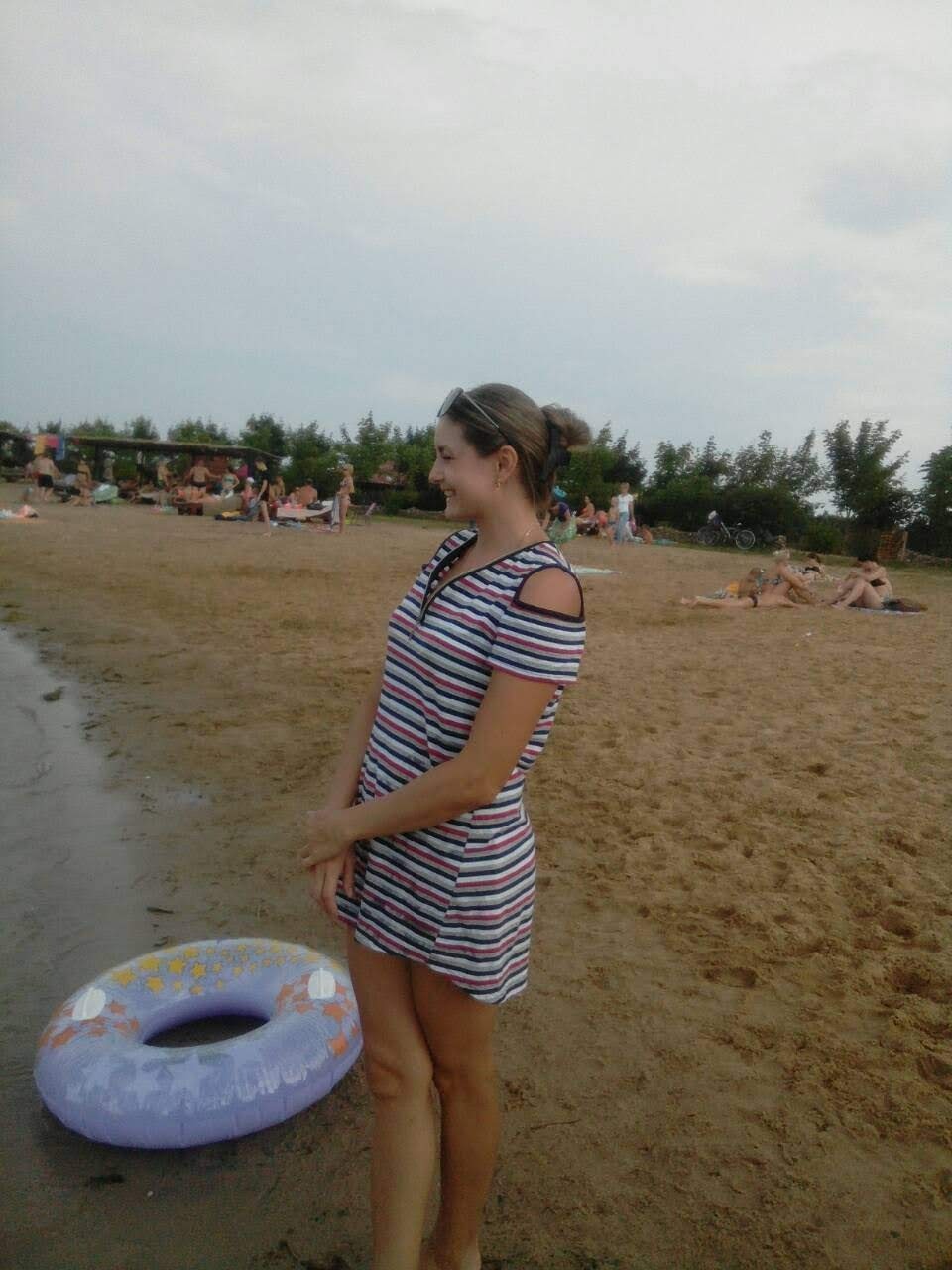 Kobieta w paski sukience stoi na piaszczystej plaży z dmuchanym kołem do pływania, w tle widać grupę osób odpoczywających.