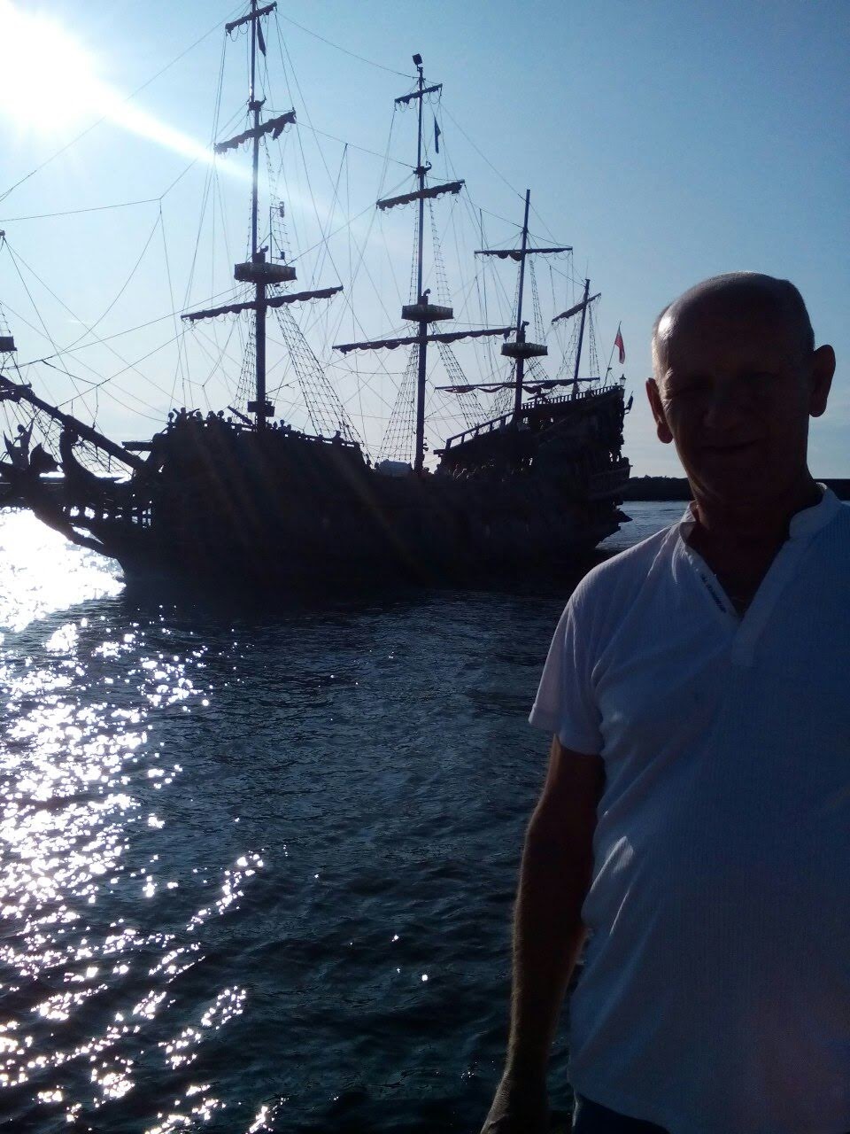 Sylwetka mężczyzny w białej koszulce na tle repliki żaglowca pirackiego na wodzie z refleksami słońca.