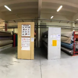 Magazyn z rolkami różnych materiałów tekstylnych, wózkiem widłowym i tablicą informacyjną o bezpieczeństwie obsługi wózka jezdniowego.