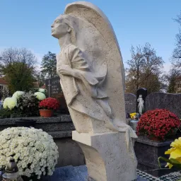 Cmentarz: Biała rzeźba anioła z rozpostartymi skrzydłami na nagrobku, otoczona kwiatami i lampionami, skierowana ku błękitnemu niebu.