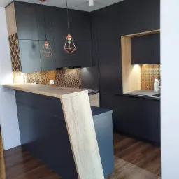 Nowoczesna kuchnia z ciemnymi szafkami, drewnianym blatem i miedzianą mozaiką na ścianie, oświetlona wiszącymi lampami w geometrycznych kloszach.