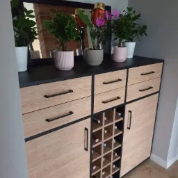 Komoda z szufladami i półkami na wino, wykończona drewnem w jasnym odcieniu i czarnymi detalami, na blacie rośliny w doniczkach i lustro odbijające ceglaną ścianę