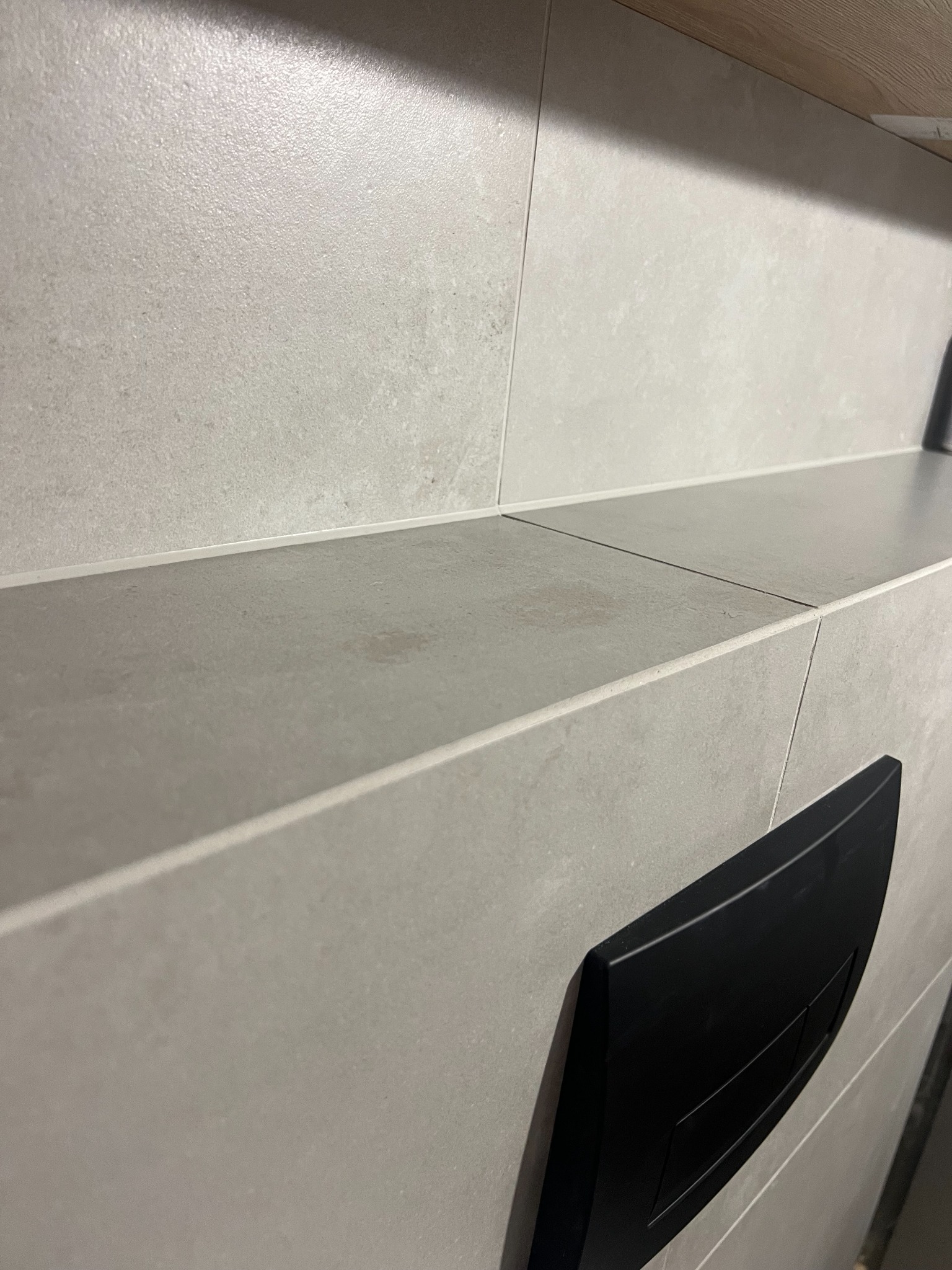 Szara, ceramiczna okładzina ścienna z półką i czarnym przyciskiem spłuczki, widoczne drobne zabrudzenia na półce.