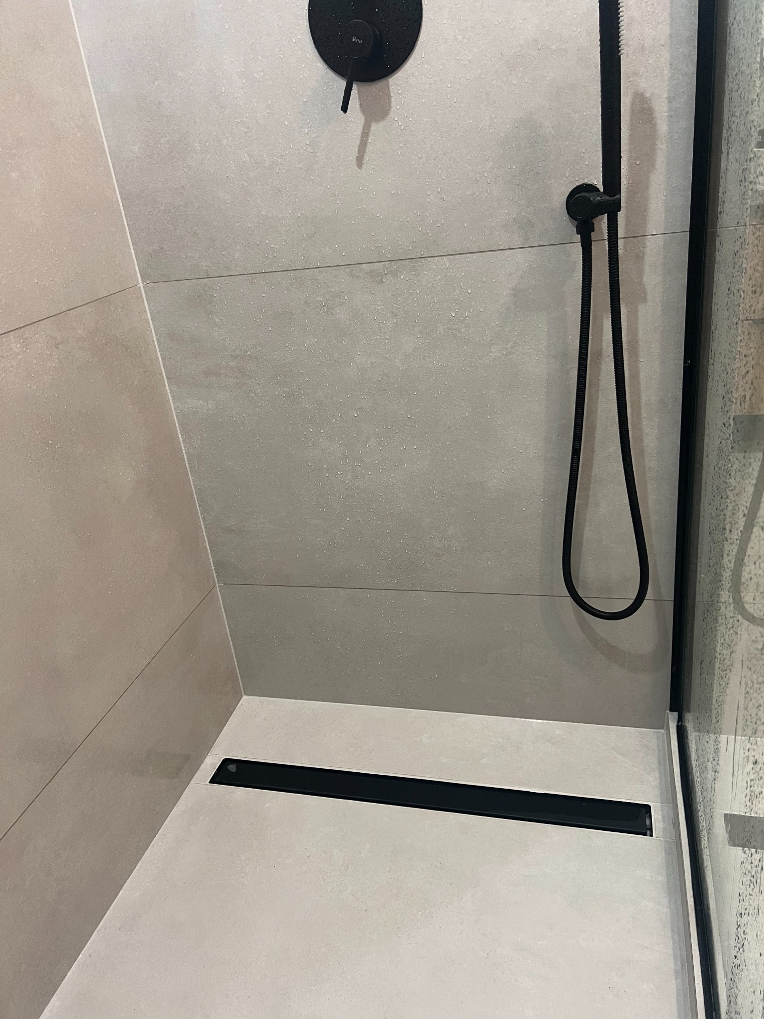 Nowoczesny prysznic z szarymi płytkami, czarną armaturą i odpływem liniowym. Na płytkach widoczne krople wody.