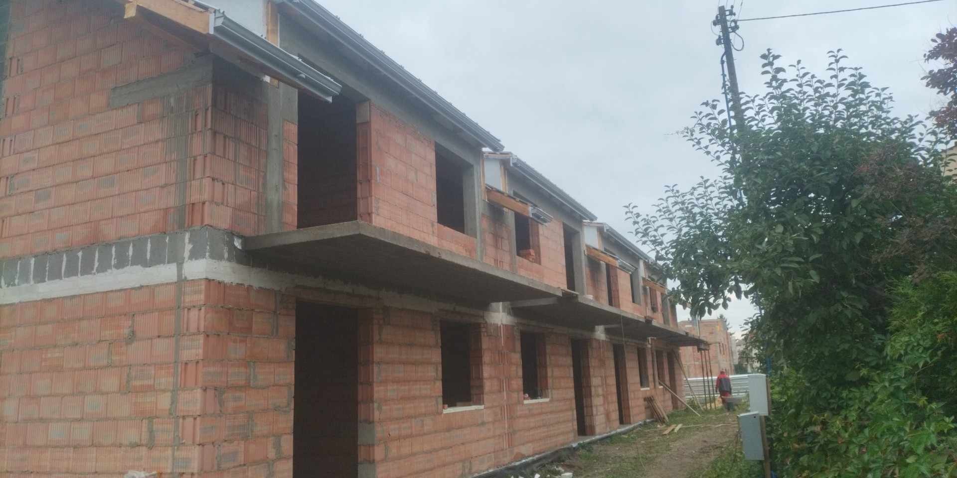 Surowy stan szeregu domów w trakcie budowy, widoczne ściany z cegły ceramicznej Porotherm, betonowe nadproża i częściowo zamontowane elementy więźby dachowej, zieleń w tle.