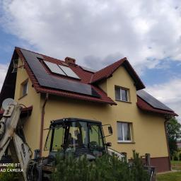 SolSerwis MIROSŁAW MAZUR - Żółty dom jednorodzinny z panelami słonecznymi na czerwonym dachu, widoczny z perspektywy ziemi, z koparką Terex 860 SX na pierwszym planie i błękitnym niebem w tle.