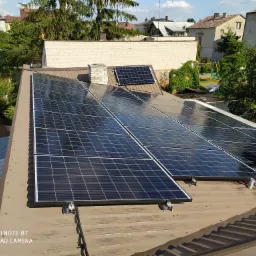 Montaż paneli słonecznych na brązowym blaszanym dachu domu jednorodzinnego; widoczny instalator w szarej czapce i rękawicach, przykręcający panel do konstrukcji wsporczej; w tle budynki mieszkalne...