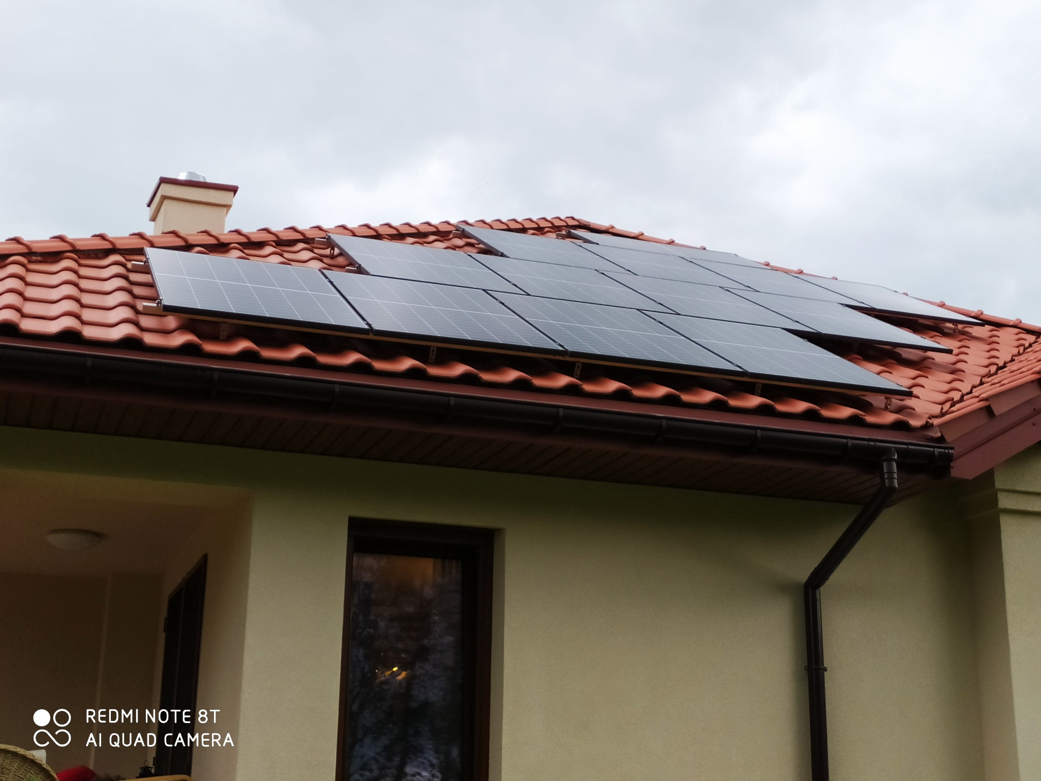 Panele słoneczne na dachu domu z czerwoną dachówką, widoczne okno i rynna spustowa.