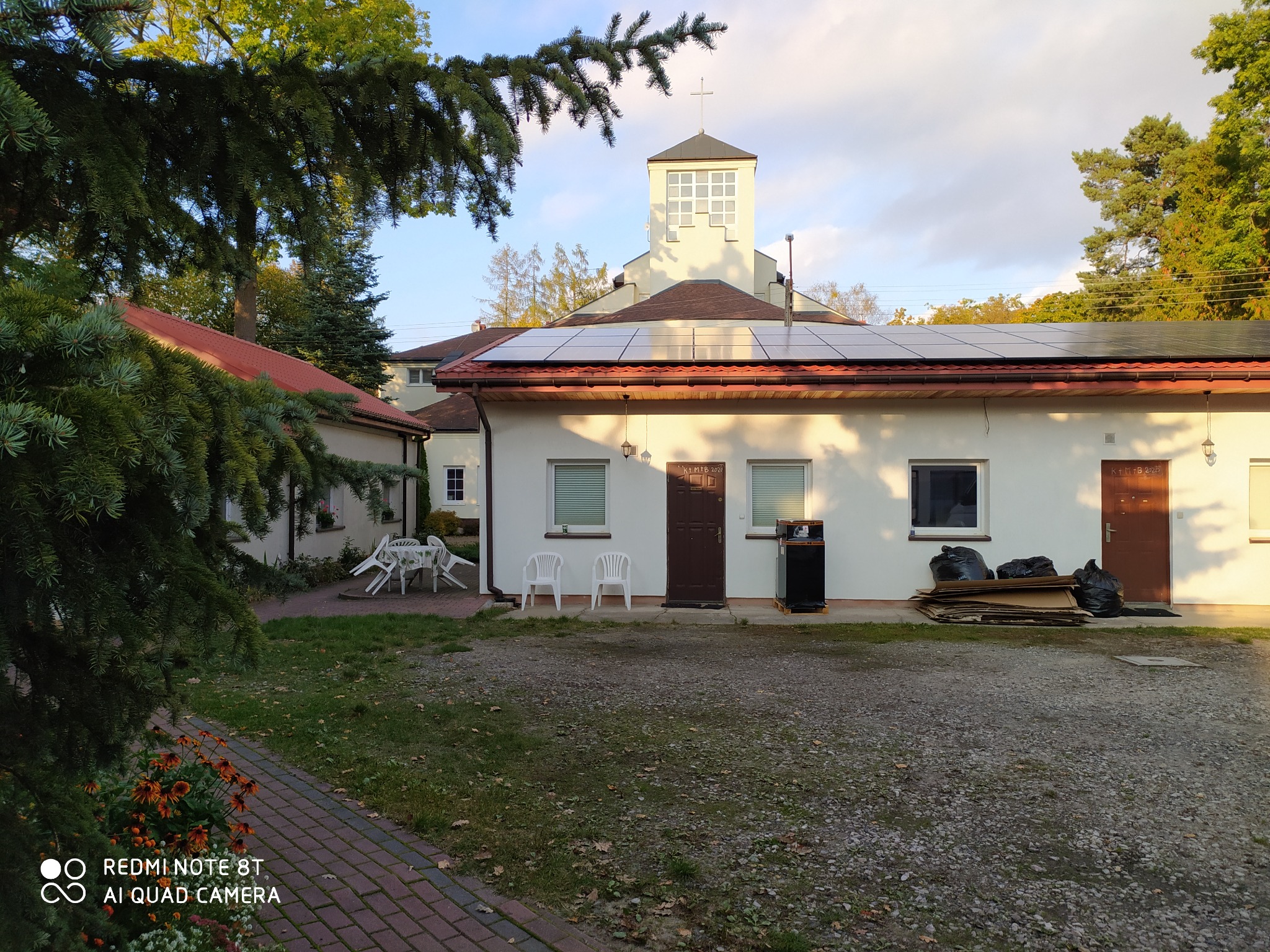 Instalacja paneli słonecznych na dachu budynku z wieżą kościelną w tle, widoczne białe ściany, brązowe drzwi oraz panele na dachu. Na ziemi leżą worki ze śmieciami i kartony.