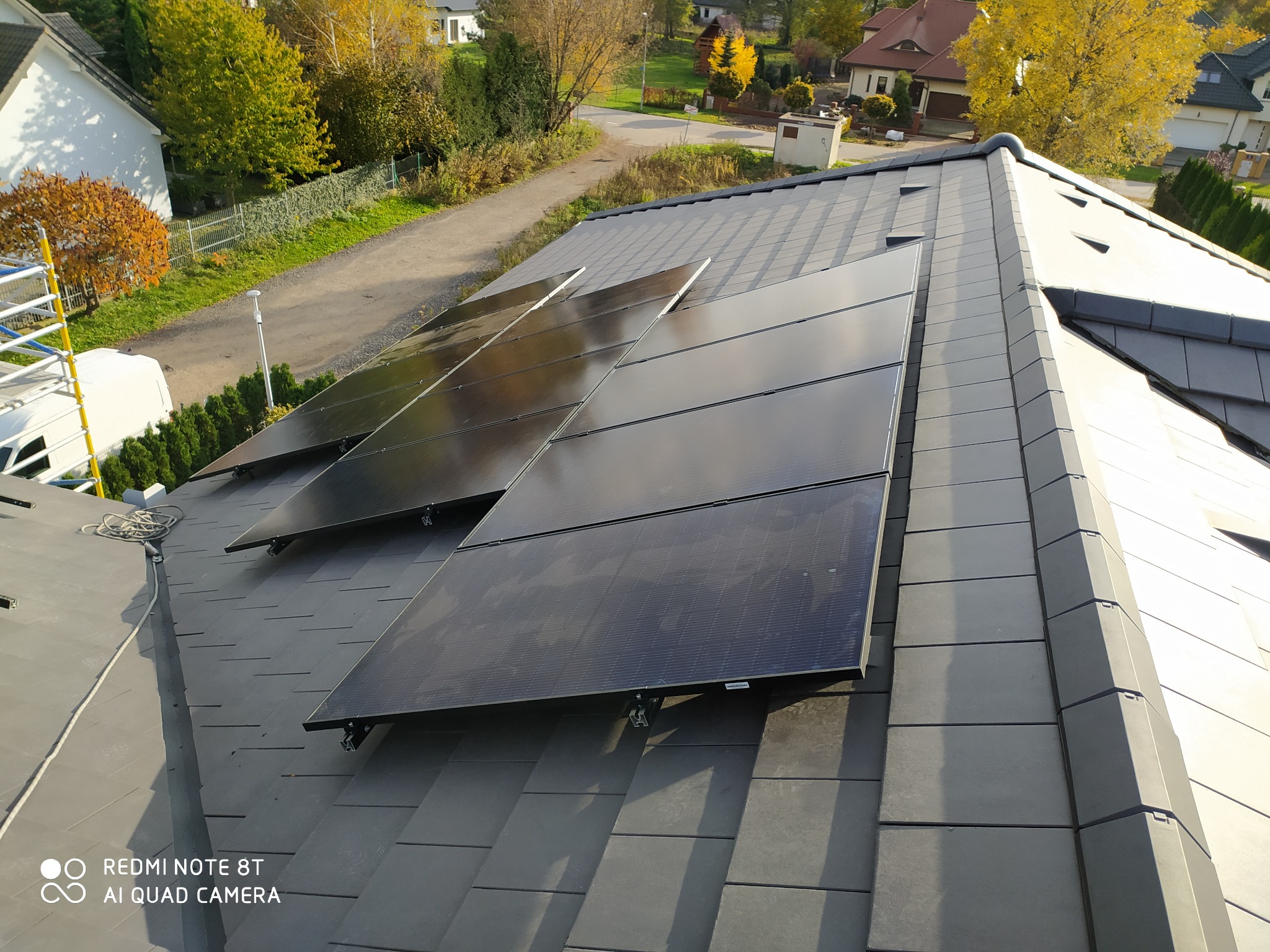 Cztery czarne panele słoneczne o łącznej mocy 3*4kWp, zamontowane na szarym dachu skośnym z dachówki ceramicznej, widok z góry na tle jesiennego krajobrazu z domami i drzewami.