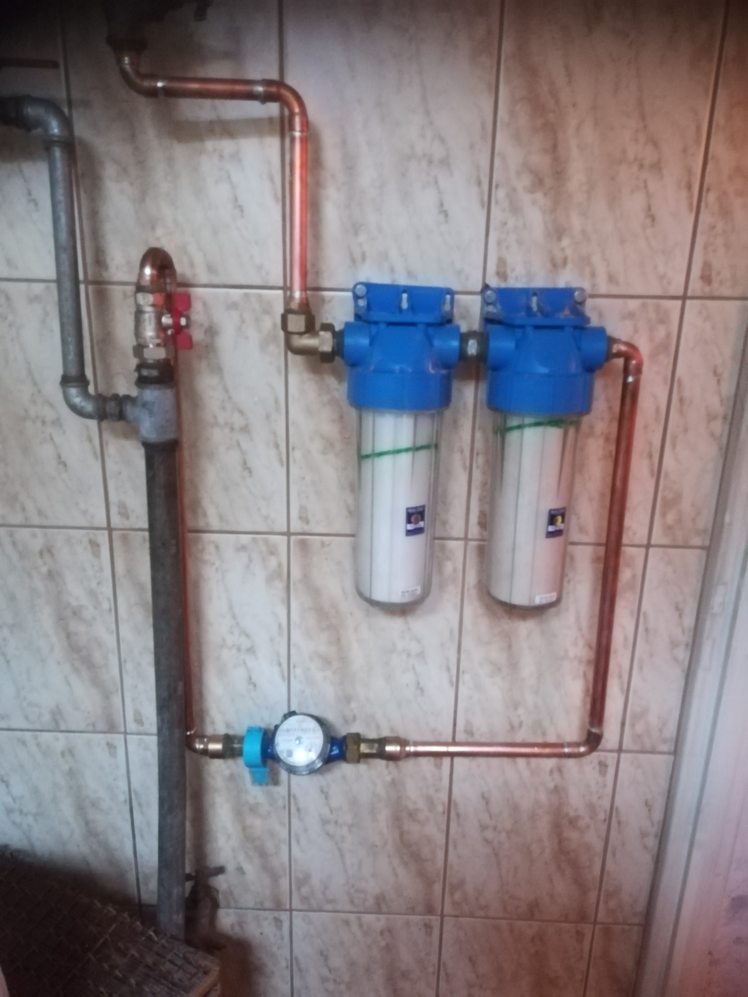 Dwa filtry wody z niebieskimi obudowami połączone miedzianymi rurami, zamontowane na ścianie z płytek ceramicznych, obok licznika wody i stalowych rur.