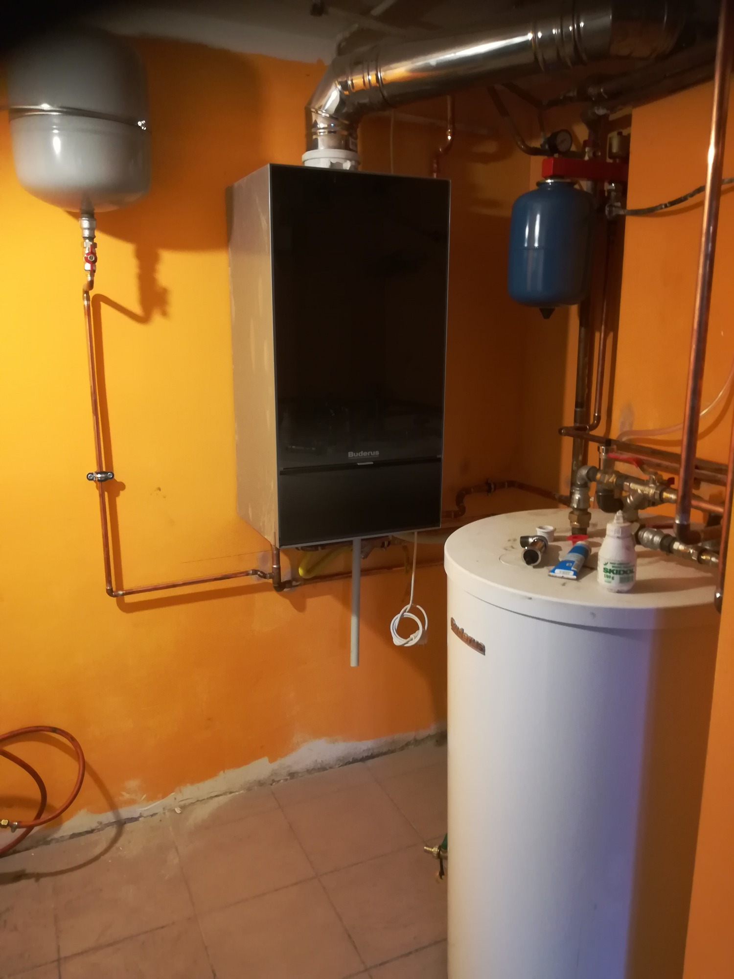 Nowoczesny piec gazowy Buderus zamontowany na pomarańczowej ścianie w piwnicy, obok biały zasobnik ciepłej wody i niebieski zbiornik wyrównawczy, widoczne miedziane rury instalacji CO i narzędzia...