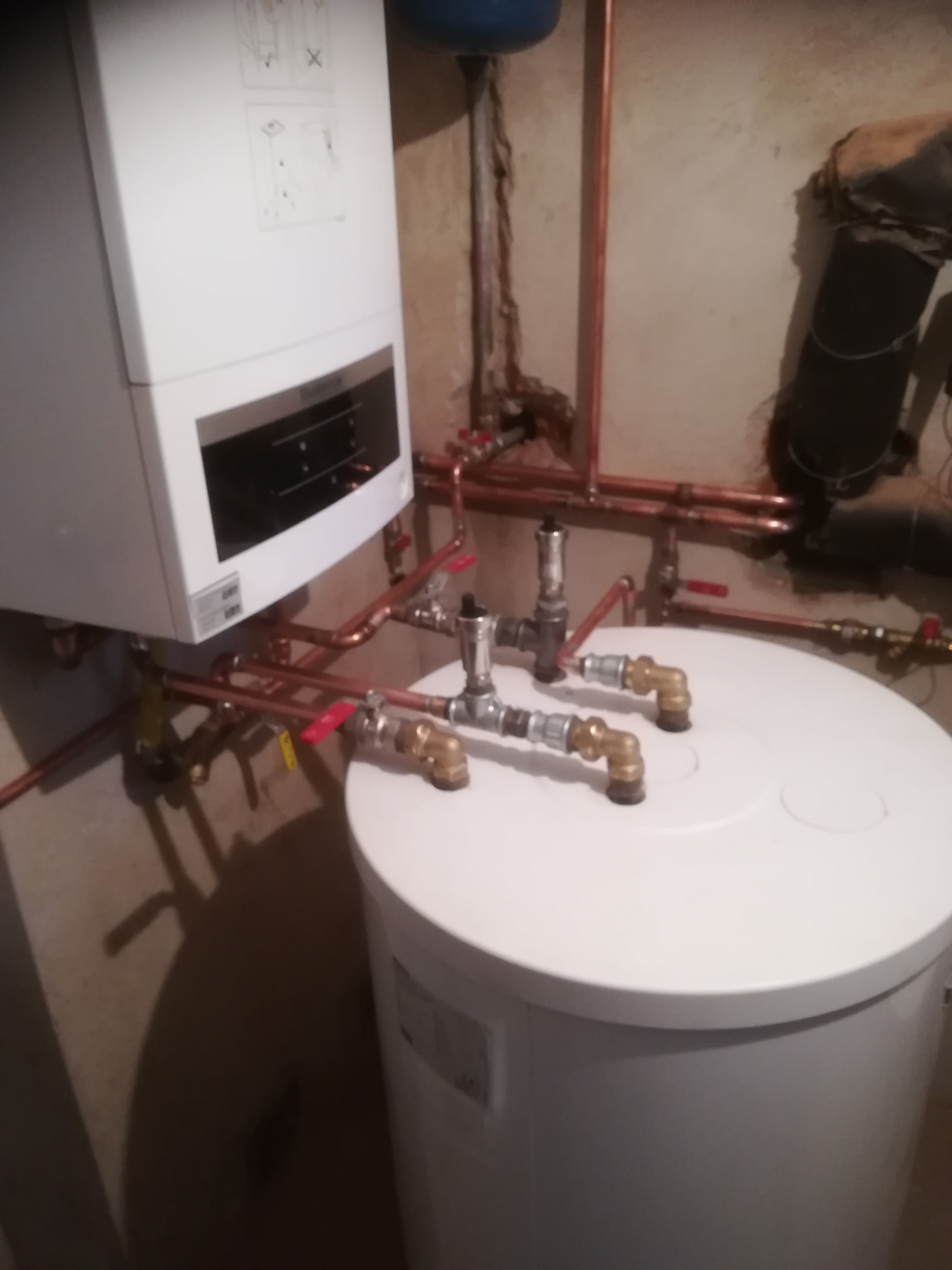 Nowoczesny piec gazowy wiszący nad białym zasobnikiem ciepłej wody, połączony miedzianymi rurami z zaworami, w surowym wnętrzu kotłowni.