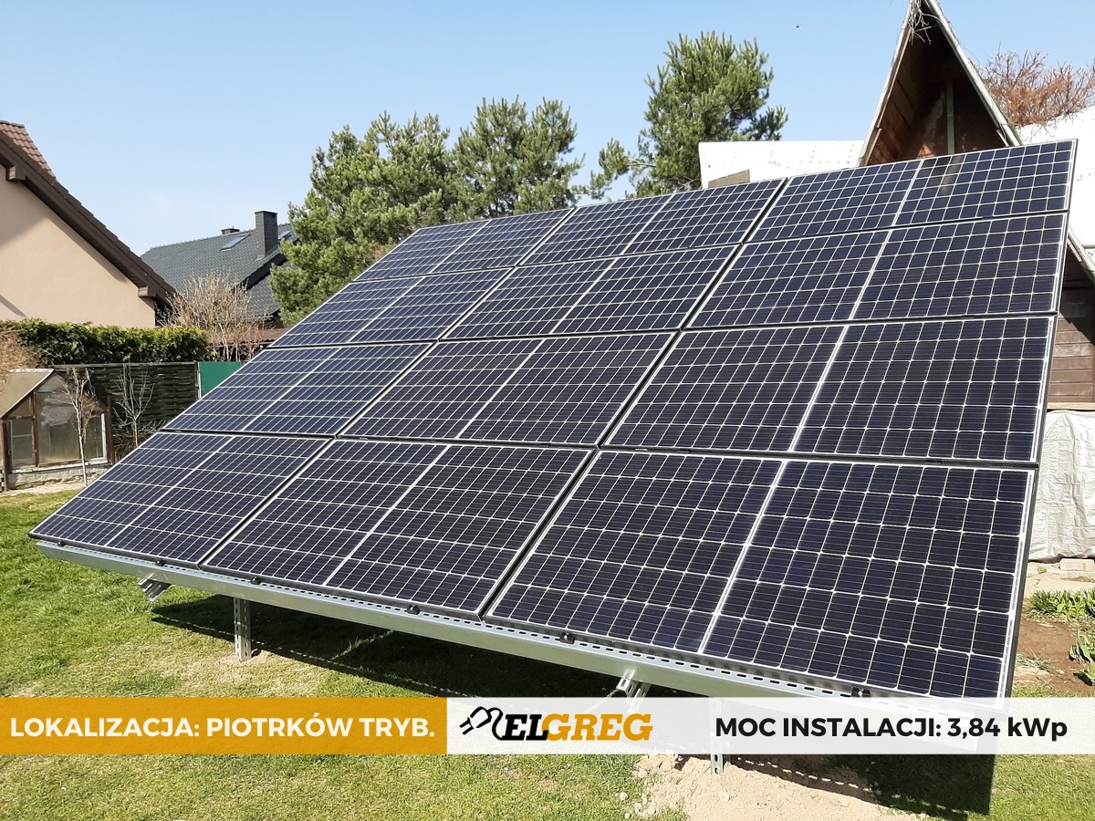 Panel fotowoltaiczny o mocy 3,84 kWp zamontowany na trawie przed domem jednorodzinnym w słoneczny dzień, widoczne elementy konstrukcyjne i logo firmy.