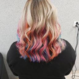 Salon fryzjerki Ewa Classic - Tył głowy kobiety z blond, falowanymi włosami z kolorowymi pasemkami w odcieniach różu, pomarańczy i błękitu, widoczne gniazdko elektryczne na ścianie.